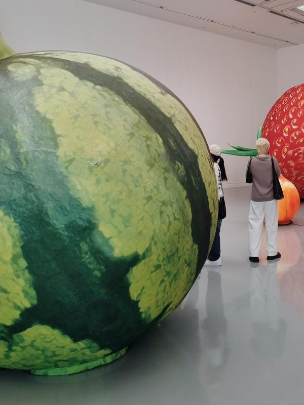 two people are standing among some giant inflated sculptures of a watermelon, an onion, and a strawberry