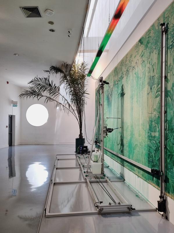 a machine paints plants on a large canvas; a palm tree sits beside it