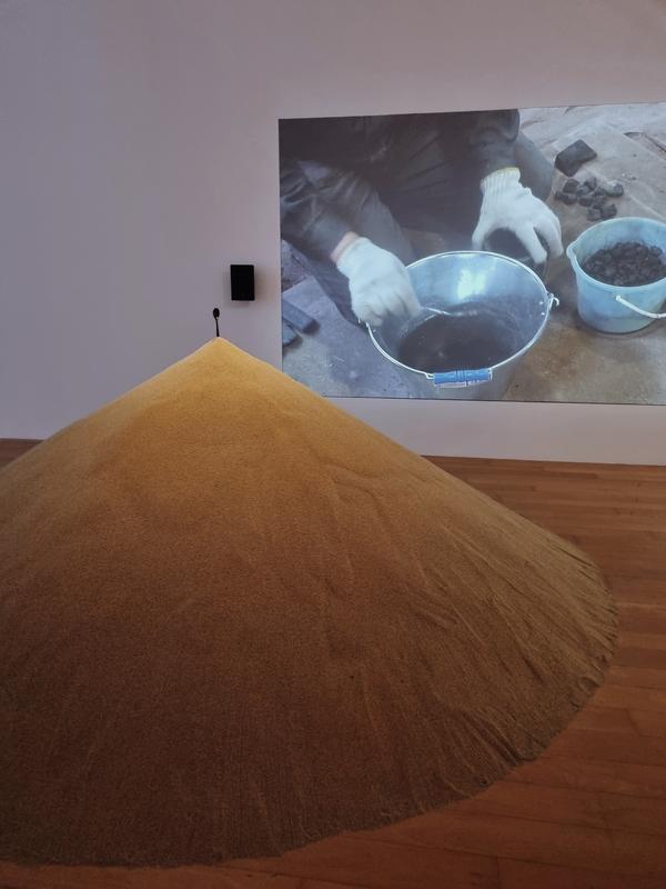 a black iron spoon sits on top of a pile of sand; a video showing the creation of said spoon is projected on the wall behind it